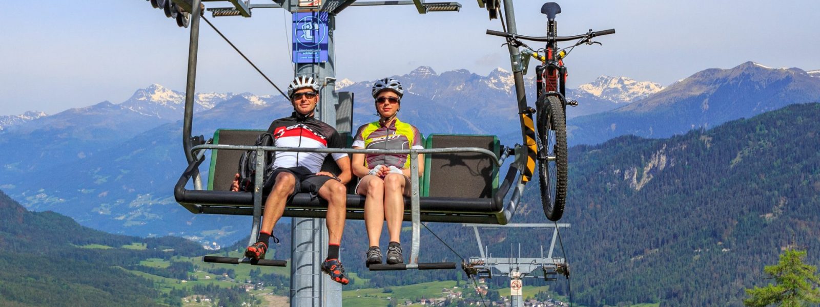 Zwei Mountainbiker fahren per Sessellift auf den Berg. Im Hintergrund der Weissensee